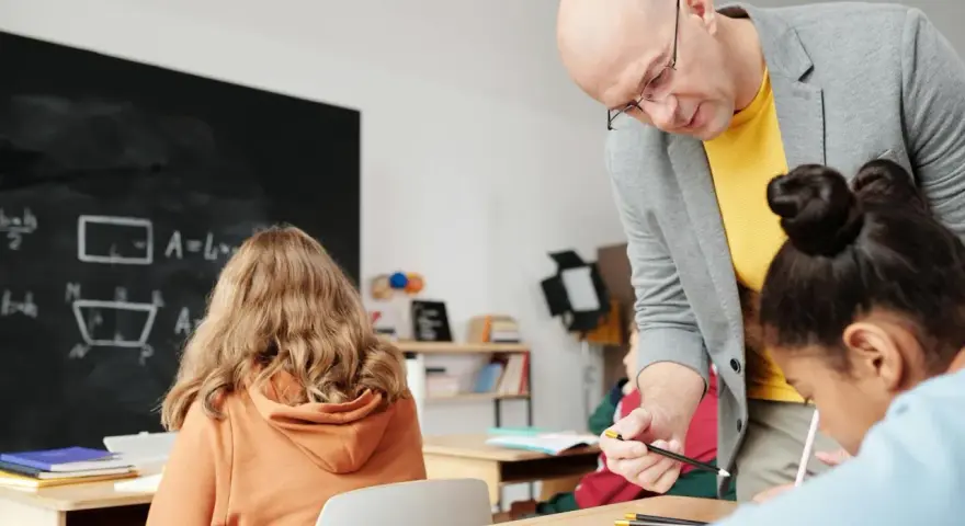 Nauczyciel pomaga uczniom podczas zajęć dydaktyczno wyrównawczych. Na tablicy wzory matematyczne.
