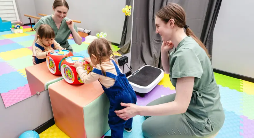 Dwie małe dziewczynki bawią się interaktywną kostką, a fizjoterapeutka wspiera ich rozwój. Www.
