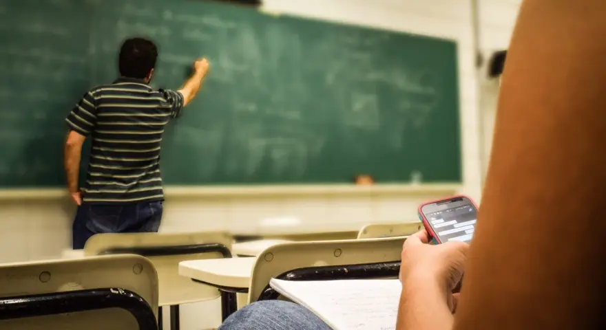 Nauczyciel pisze na tablicy, a uczeń w pierwszej ławce przegląda telefon. Obowiązki nauczyciela to nie tylko nauczanie, ale też pilnowanie porządku.