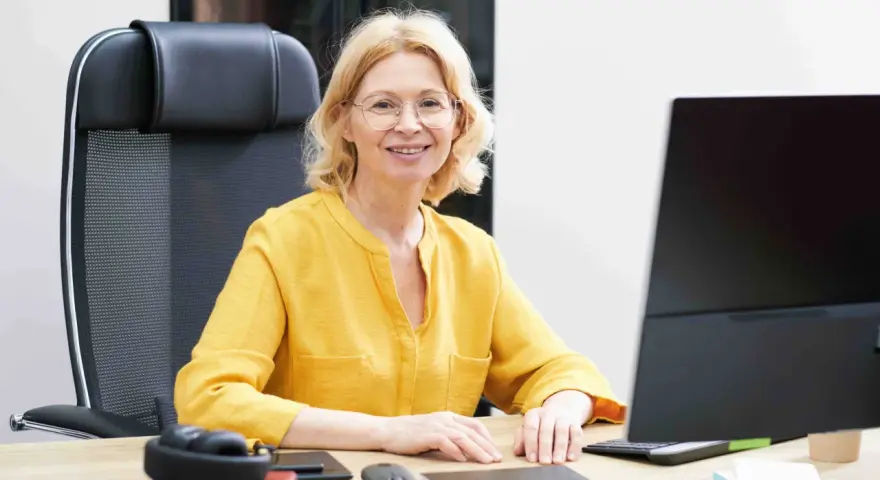 Uśmiechnięta wicedyrektor w okularach siedzi przy biurku, przed nią komputer i słuchawki.