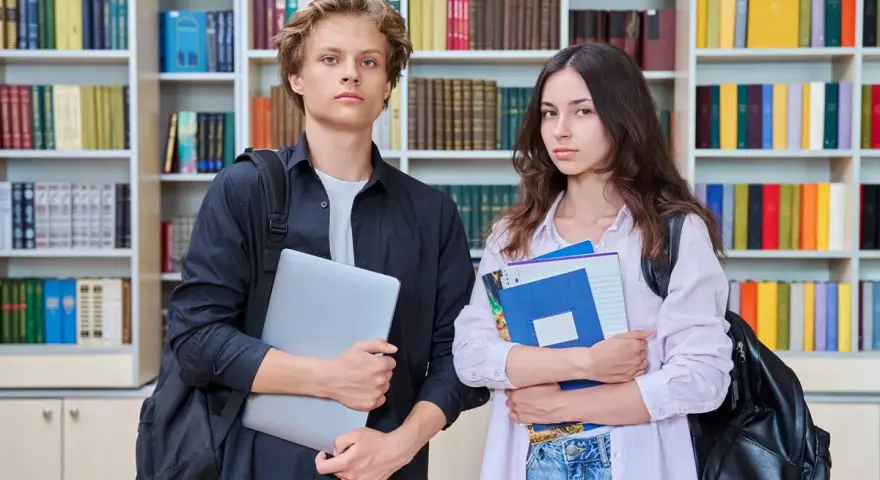 Dwóch studentów z książkami i laptopem na tle regałów z książkami.