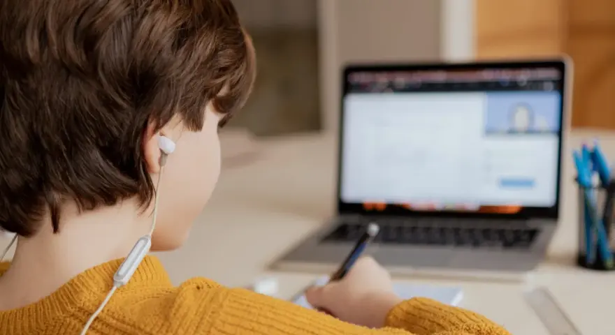 Chłopiec w żółtym swetrze uczy się zdalnie, pisząc notatki. Czy uczniowie dostaną laptopy, by móc tak pracować?