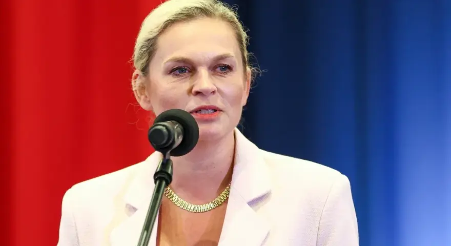 Kobieta w jasnej marynarce mówi do mikrofonu na tle czerwono-niebieskiego tła.