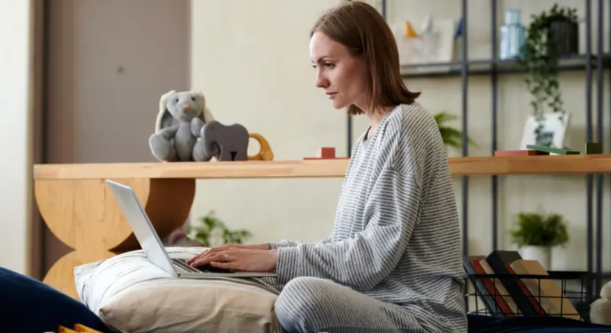 Kobieta na łóżku pisze wniosek o urlop rodzicielski na laptopie, otoczona zabawkami i pluszakami.