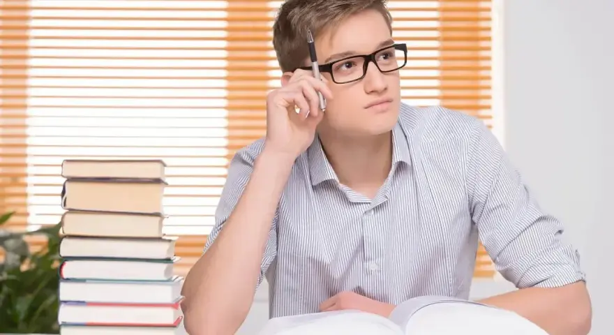Młody mężczyzna w okularach, z długopisem w dłoni, zamyślony nad książką. Stos książek obok. Jego statut to student.