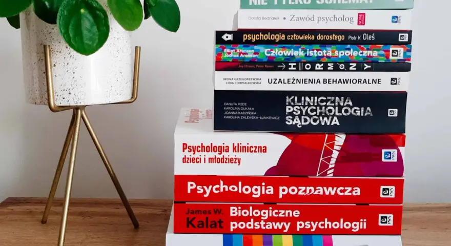 Stos książek o psychologii, od ogólnych po specjalistyczne, sugeruje, ile trwają studia psychologiczne. Obok zielona roślina.
