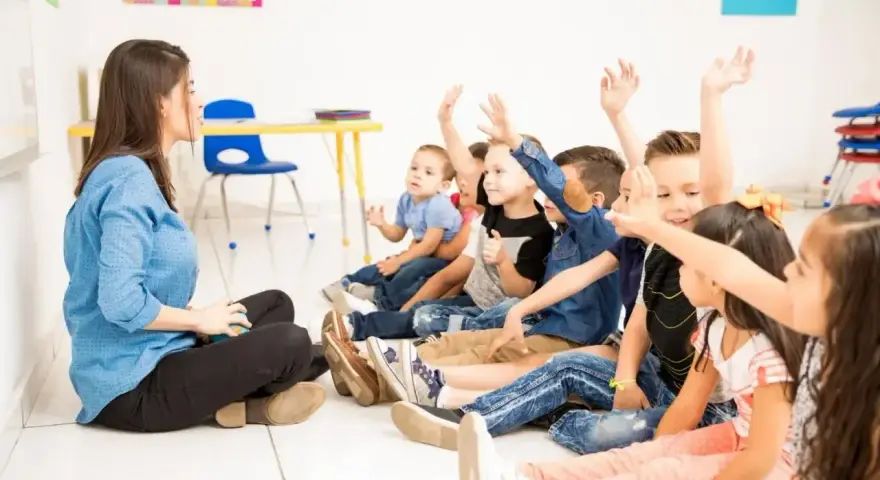 Nauczycielka siedzi na podłodze, a dzieci z podniesionymi rękami odpowiadają na pytania. Widać, że frekwencja w szkole jest wysoka, a uczniowie chętnie uczestniczą w lekcji.