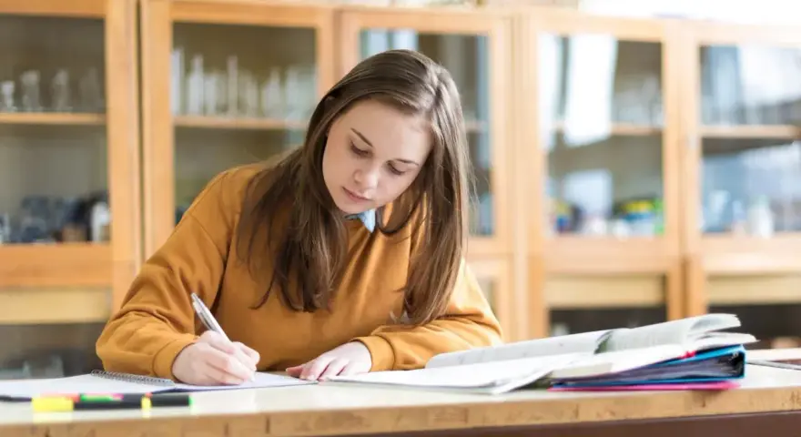Dziewczyna pisze w zeszycie, przygotowując się do egzaminu. Marzy o stypendium szkolnym, które pomoże jej w dalszej nauce.