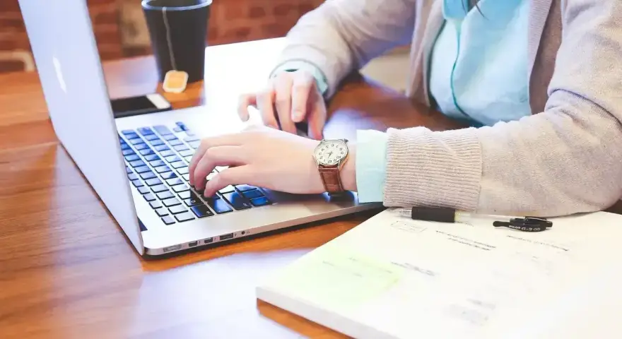 Osoba pisząca na laptopie, z kubkiem herbaty i notatkami na biurku.