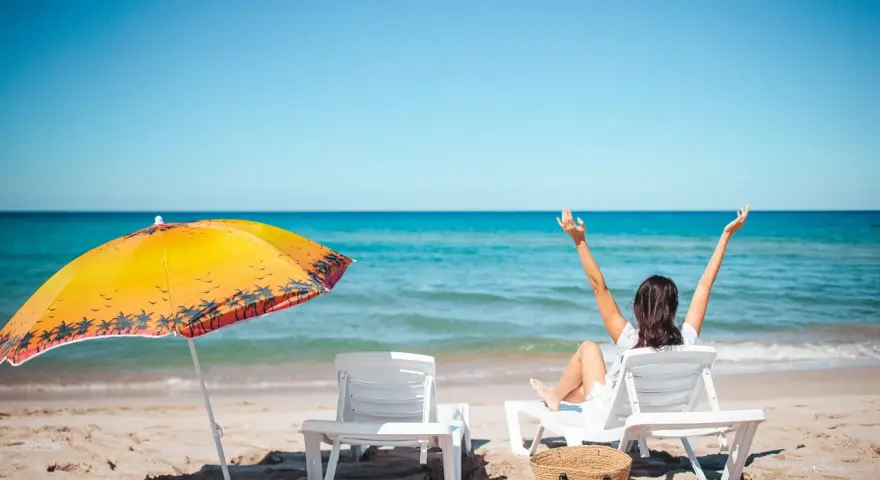 Kobieta cieszy się słońcem na plaży, w pełni wykorzystując swój urlop. Obok parasol i leżaki.