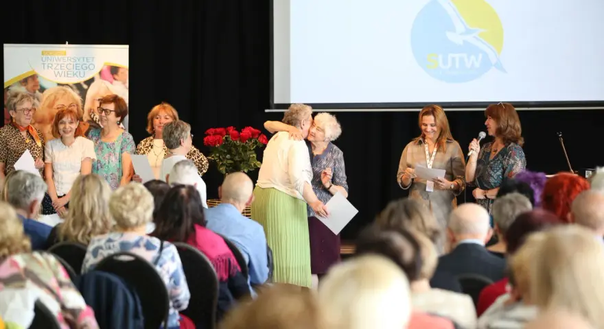 Uroczystość w Sopockim Uniwersytecie Trzeciego Wieku. Uśmiechnięci seniorzy, wręczanie dyplomów, wystąpienie.
