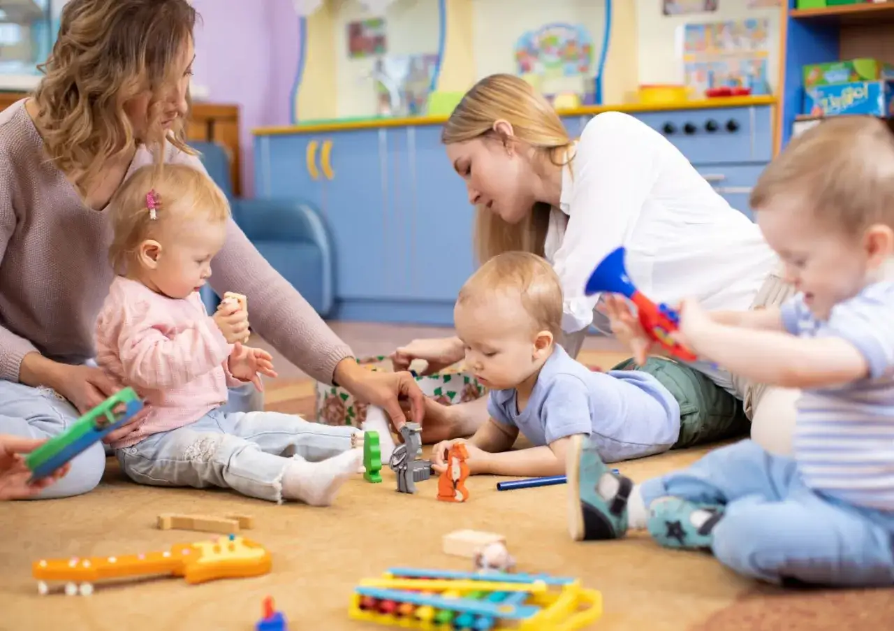 Dwie opiekunki dziecięce bawią się z maluchami na dywanie, otoczone kolorowymi zabawkami.