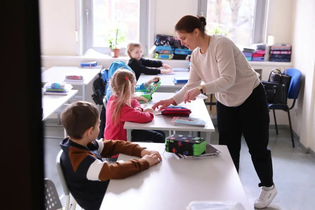 Nauczycielka pomaga uczniom w lekcji, przypominając o obowiązku szkolnym. Dzieci siedzą przy ławkach, skupione na zadaniach.