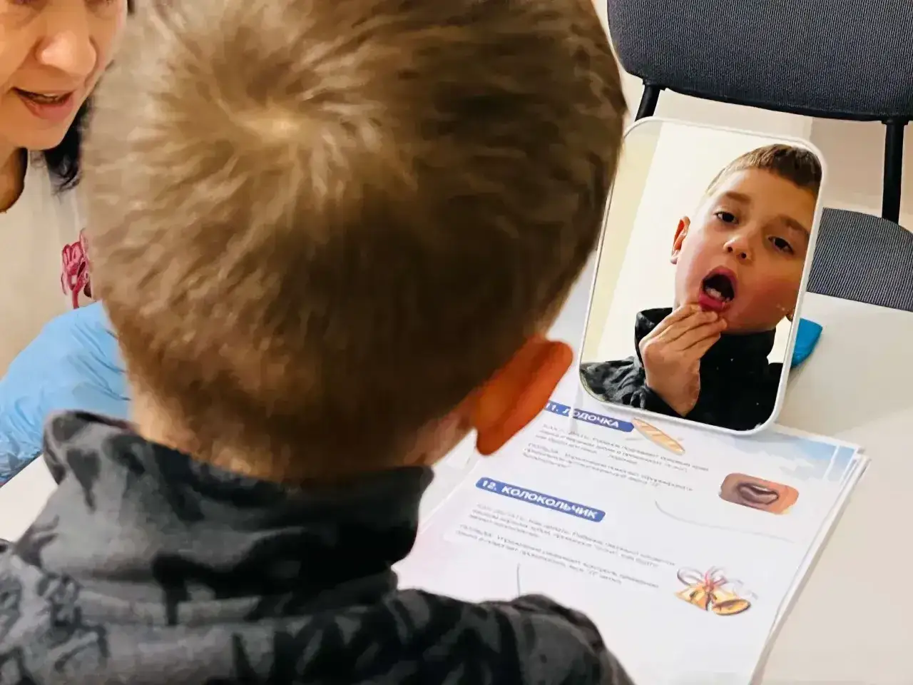 Chłopiec z niepełnosprawnością intelektualną w stopniu umiarkowanym ćwiczy artykulację przed lustrem, pod okiem terapeuty.
