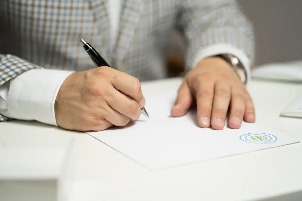 Mężczyzna w garniturze podpisuje umowę na zastępstwo. Dłoń z długopisem nad białą kartką papieru.