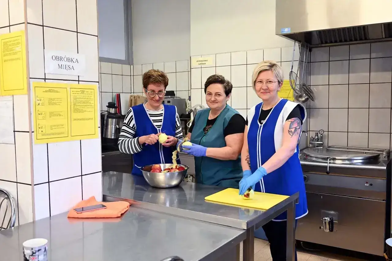 Trzy kobiety w fartuchach przygotowują jabłka w kuchni. Pracują w zespole, jak sprawny intendent.
