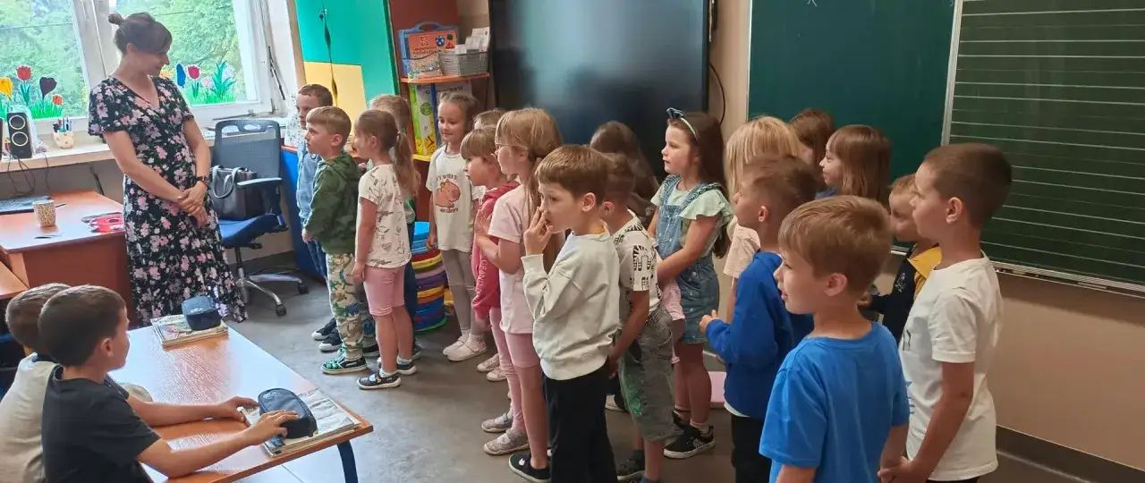 Nauczycielka w kwiecistej sukience stoi przed grupą dzieci w oddziale przedszkolnym, które słuchają jej uważnie.