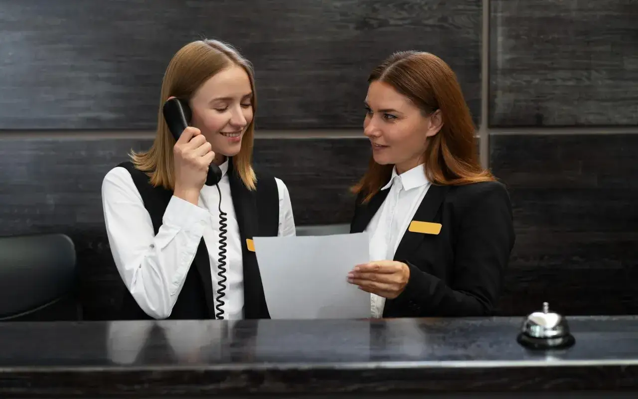 Dwie pracownice recepcji, jedna rozmawia przez telefon, druga trzyma dokument. Woźny właśnie przyniósł im kawę.
