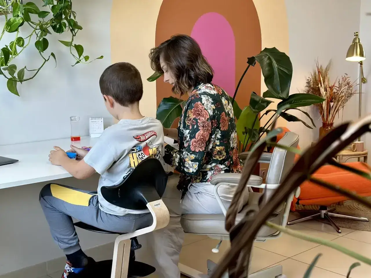 Chłopiec siedzi przy biurku, a kobieta pochyla się nad nim, wspierając go w procesie diagnozy psychologicznej.