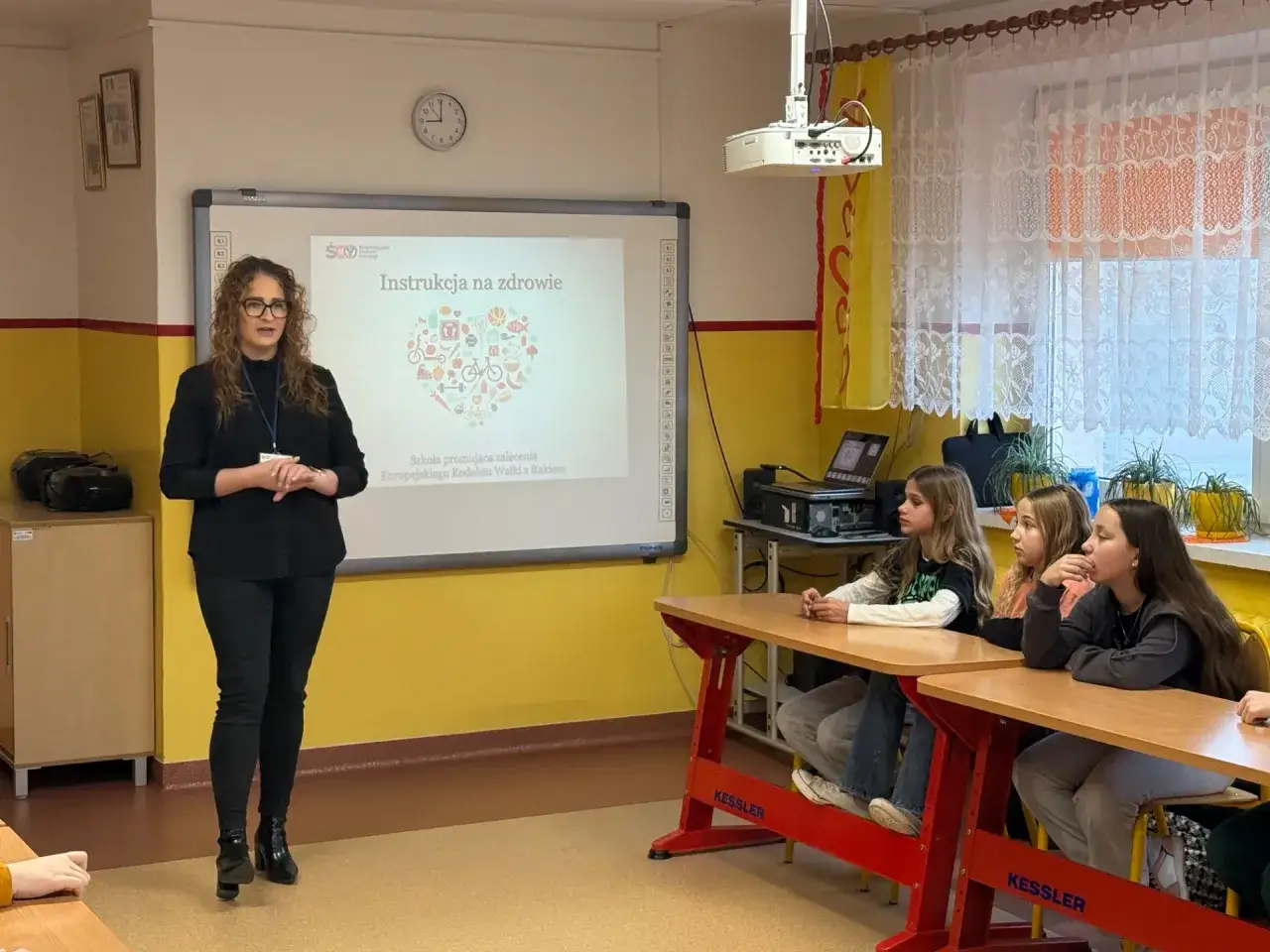 Nauczycielka prowadzi lekcję o zdrowiu, prezentując na tablicy serce z symbolami zdrowego stylu życia. To część programu nauczania edukacja zdrowotna.