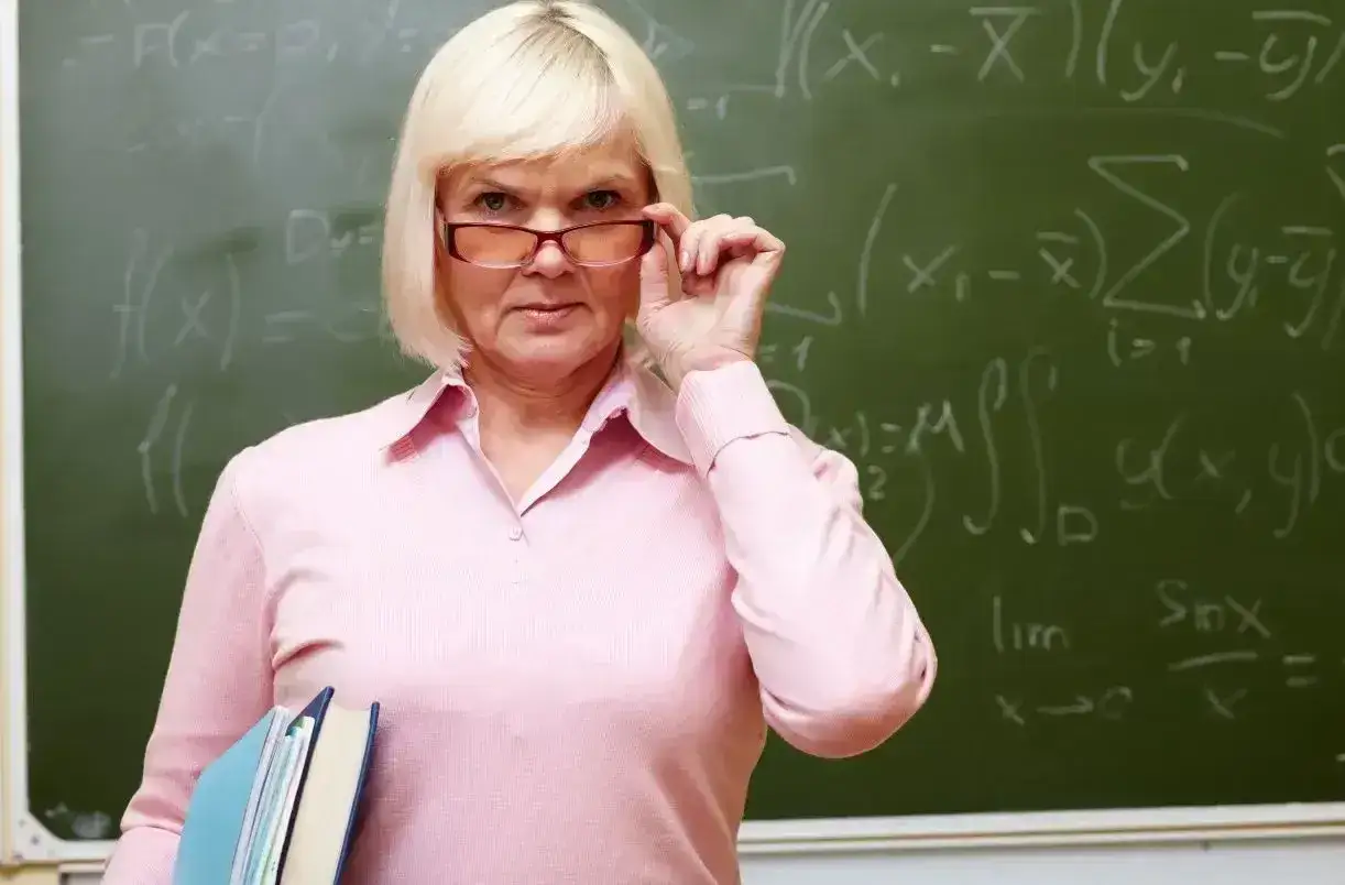 Nauczycielka matematyki z okularami i książkami, na tablicy wzory. Jej wiedza to świadczenie kompensacyjne dla uczniów.