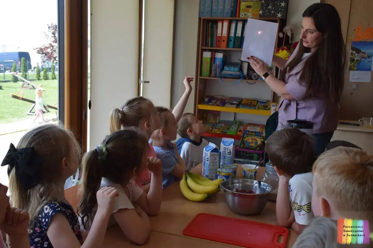 Dzieci słuchają nauczycielki podczas zajęć o zdrowym odżywianiu. Na stole owoce i jogurty.