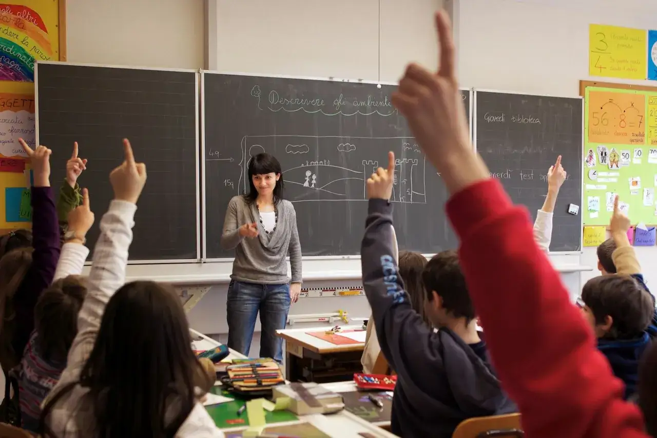 Nauczycielka prowadzi zajęcia dydaktyczno wyrównawcze. Uczniowie z podniesionymi rękami aktywnie uczestniczą w lekcji.