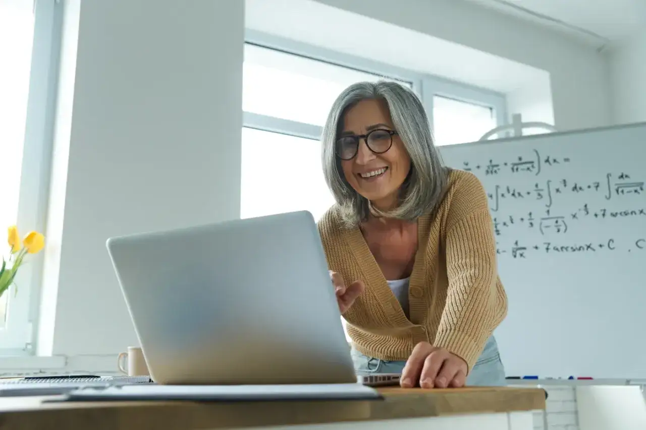 Uśmiechnięta nauczycielka z siwymi włosami, pracująca przy laptopie. W tle tablica z równaniami matematycznymi, symbolizująca jej doświadczenie i przyszłe emerytury dla nauczycieli.