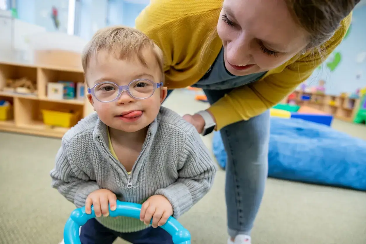 Mały chłopiec z zespołem Downa, w okularach, z językiem na brodzie, bawi się z uśmiechniętą oligofrenopedagog w sali przedszkolnej.