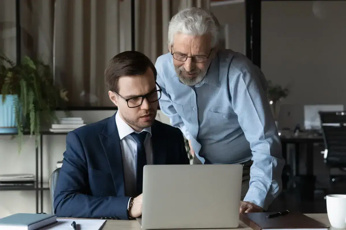 Dwóch mężczyzn analizuje dane na laptopie. Starszy doradza młodszemu, jak najlepiej obliczyć dodatek za wysługę lat w budżetówce.