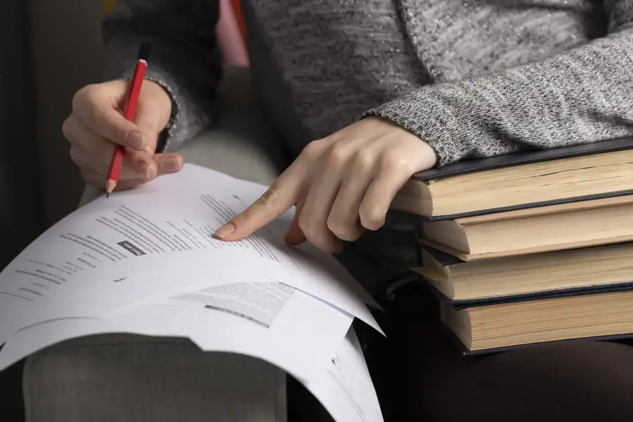 Ręka studenta z ołówkiem wskazuje na dokument, stos książek obok. Czekanie na wyniki matur.