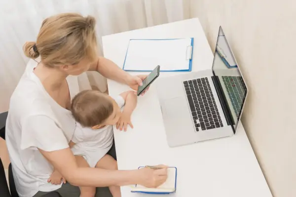 Kobieta z dzieckiem na kolanach sprawdza na telefonie informacje o zasiłku macierzyńskim 80 czy 100% i 60. Obok laptop i notatnik.