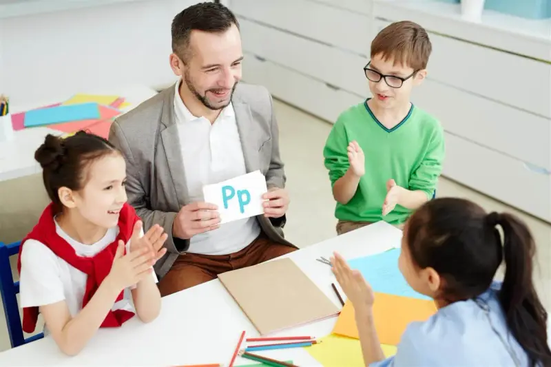 Nauczyciel pokazuje literę "Pp" podczas zebrania z rodzicami. Dzieci klaszczą, ucząc się alfabetu.