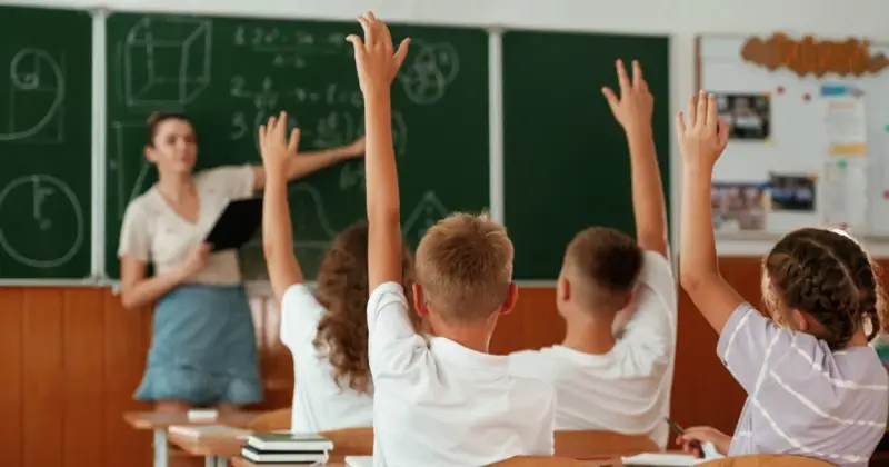 Uczniowie z podniesionymi rękami na lekcji matematyki. Nauczycielka przy tablicy, na której widać wzory. Wszyscy chętni do odpowiedzi.