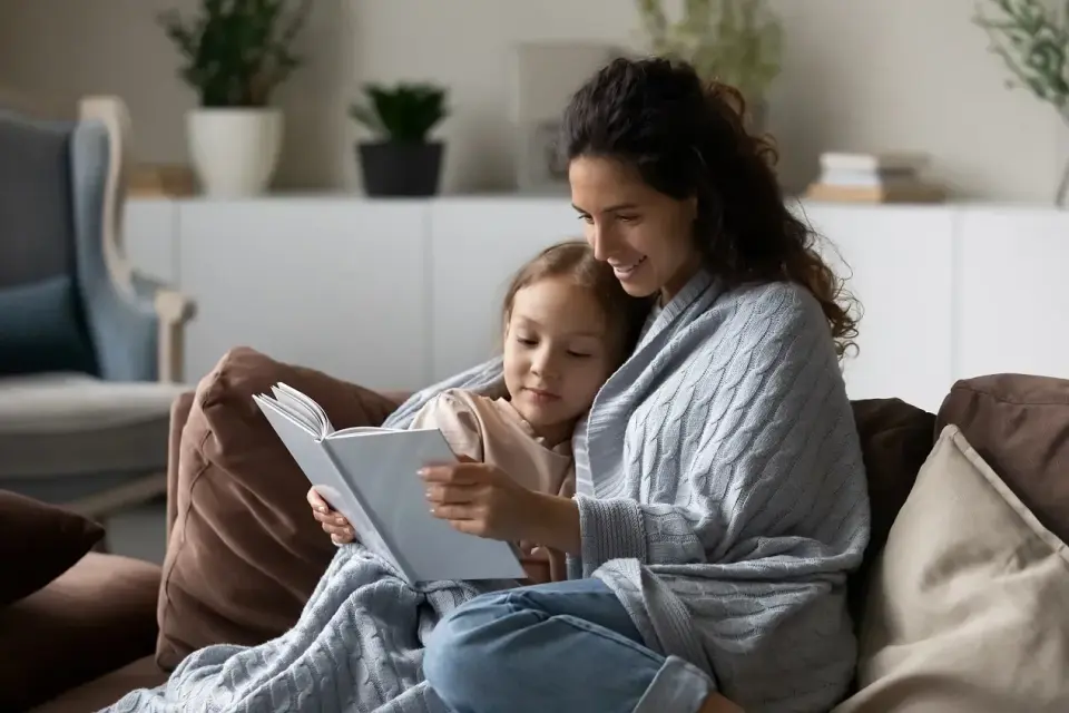 Ciepła opieka na dziecko 2 dni. Mama czyta bajkę córeczce, otulone kocem na kanapie.